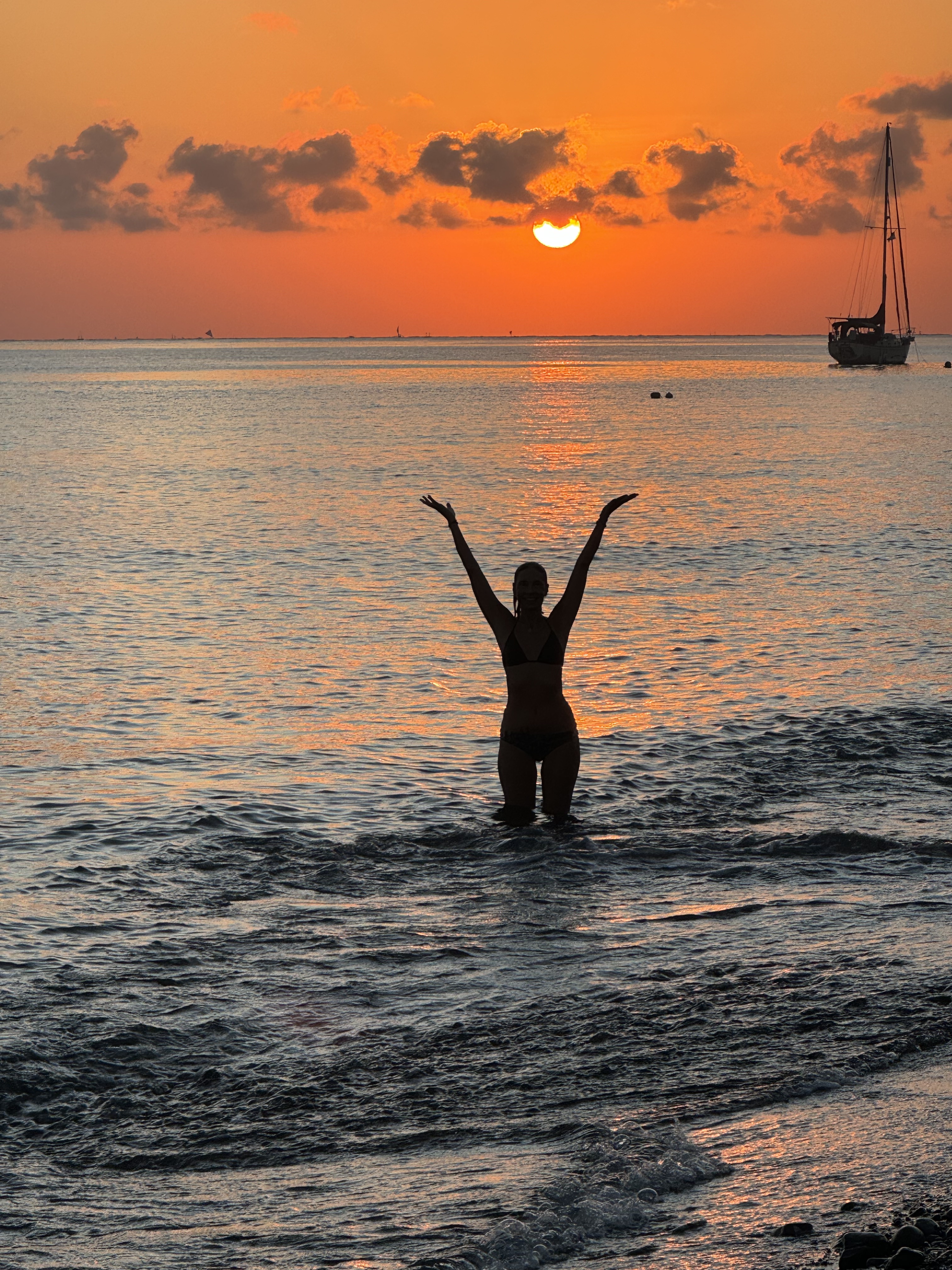 Silhouette of a person standing in the ocean at sunset with arms raised, with a sailboat in the background.